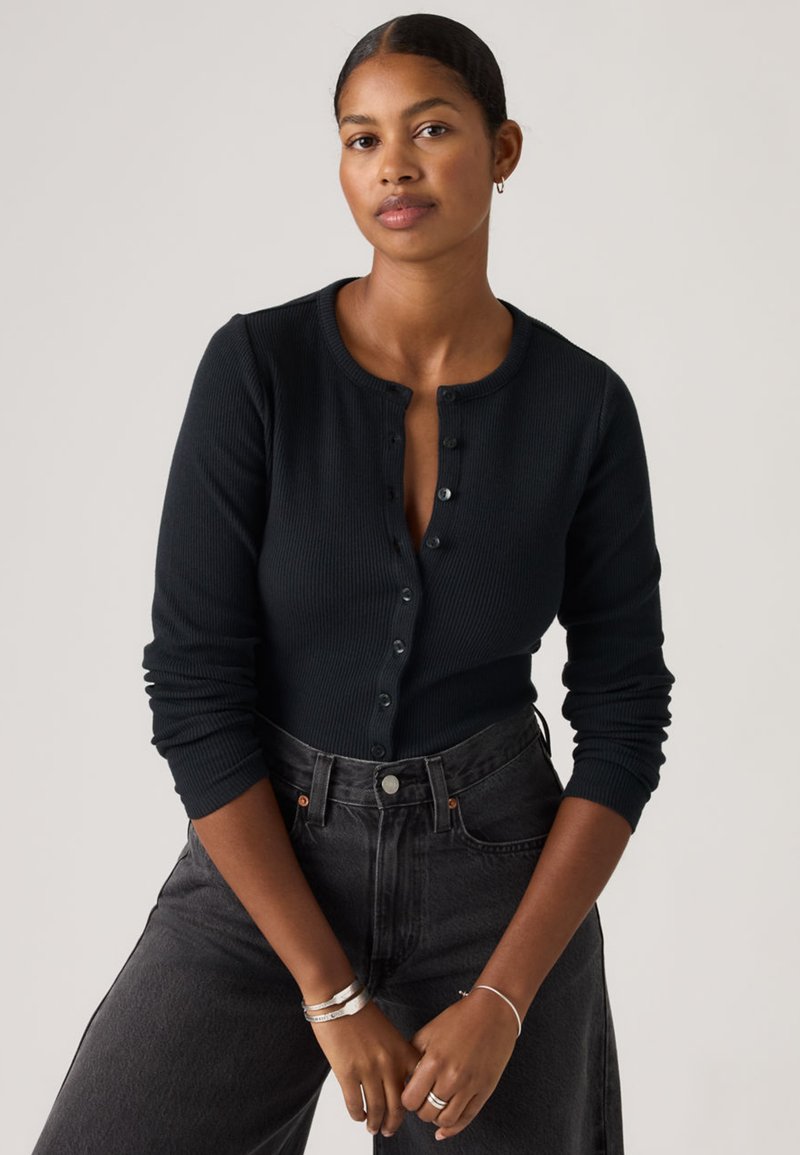 Cárdigan negro de manga larga con tejido acanalado, escote redondo, cierre frontal con botones y puños ajustados. Combinado con vaqueros oscuros de cintura alta.