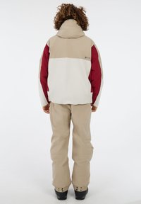 Sneeuwjack in wit, beige en bordeauxrood met een capuchon; heeft een kleurblokontwerp en elastische manchetten. Beige broek en zwarte laarzen.