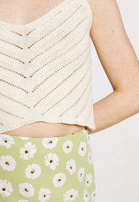 Top corto de punto en color crema con un patrón de chevron. Combinado con una falda verde con estampados florales blancos. Textura suave y diseño ajustado.