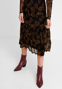 Kvinna som bär en svart midikjol med orange blommönster och burgunderfärgade spetsiga ankelstövlar på en vit bakgrund.