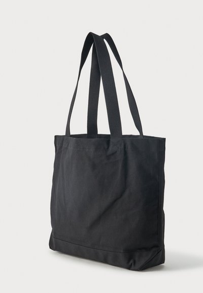 Μαύρη υφασμάτινη τσάντα tote με μακριές ιμάντες ώμου, όρθια σε φωτεινό φόντο.