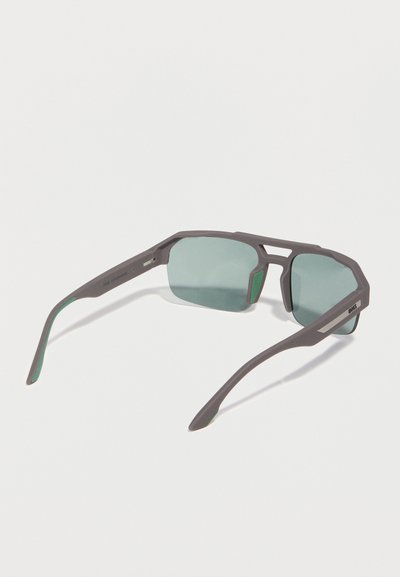 Lunettes de sport grises avec des verres rectangulaires teintés verts et des montures angulaires, vues de dos sur un fond blanc.