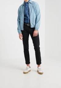 Ljusblå jeansjacka, mönstrad marinblå skjorta, svarta byxor och vita sneakers med bruna sulor. Jackan har elastiska ärmslut och krage.