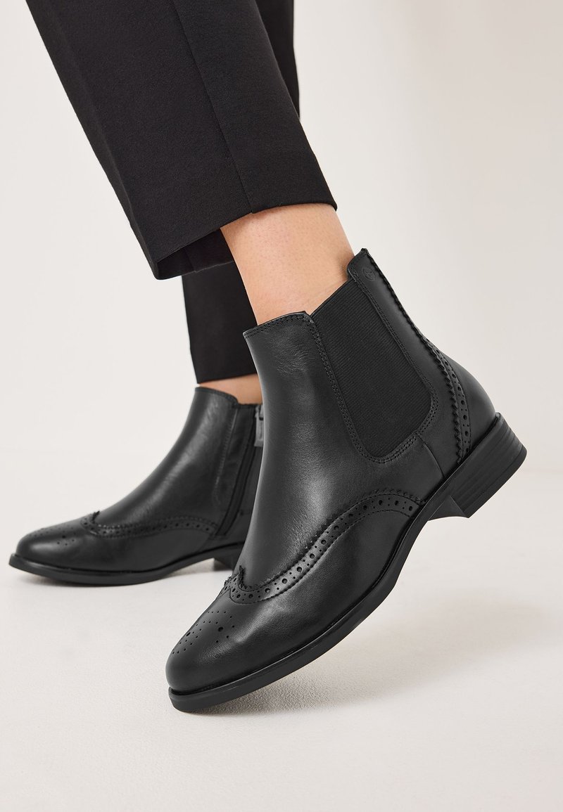Svarta läderankelbooties med elastiska sidopaneler, spetsig tå och dekorativ perforerad broguedetalj. Låg staplad klack.