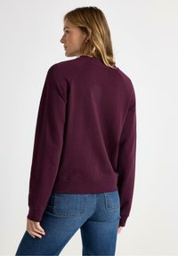 Bordeaux sweatshirt med ribbestrikket krave og manchetter, lavet af blødt stof, med raglanærmer, parret med blå jeans.