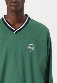 Cămașă verde cu mâneci lungi, dintr-un material ușor, cu decolteu în formă de V, având o margine în dungi albastre și albe și un logo alb pe partea stângă a pieptului.