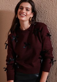 Burgunderfarbener gestrickter Pullover mit schwarzen Satinschleifen an Ärmeln und Vorderseite, rundem Ausschnitt und gerippten Bündchen.