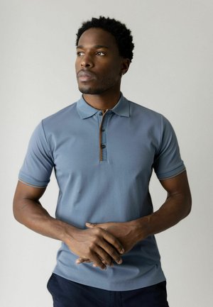 Ung mand med kort krøllet hår iført en blå poloshirt, der ser tænksomt til siden med hænderne foldet foran sig.