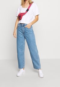 Bílý top s balónovými rukávy, světle modré džíny s vysokým pasem a bílé tenisky. Křížem přes tělo je nošena ledvinka s růžovým popruhem.