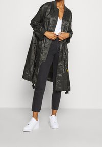 Lång, mörkgrön regnkappa med en glänsande yta, tryckknappsstängning och sidofickor, bärs över en vit topp och svarta jeans, tillsammans med vita sneakers.