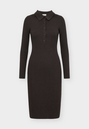 Femme portant une robe en maille côtelée noire ajustée, manches longues, col boutonné, et des bottines noires, debout devant un fond uni.
