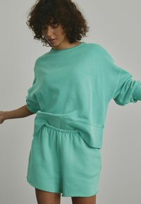 Un hanorac oversized de culoare turcoaz deschis, asortat cu pantaloni scurți corespunzători. Hanoracul are mâneci largi, iar pantaloni scurți au un tiv elastic.