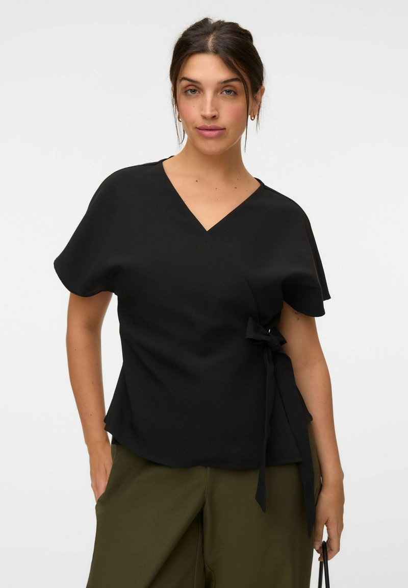 Femme aux cheveux foncés attachés porte un chemisier cache-cœur noir avec un lien sur le côté et un pantalon vert olive, debout sur un fond blanc uni.