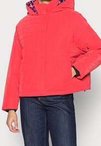 Rød quiltet jakke med foret hætte, der har et pink mønstret indre, lange ærmer og frontlommer, parret med mørkeblå jeans.