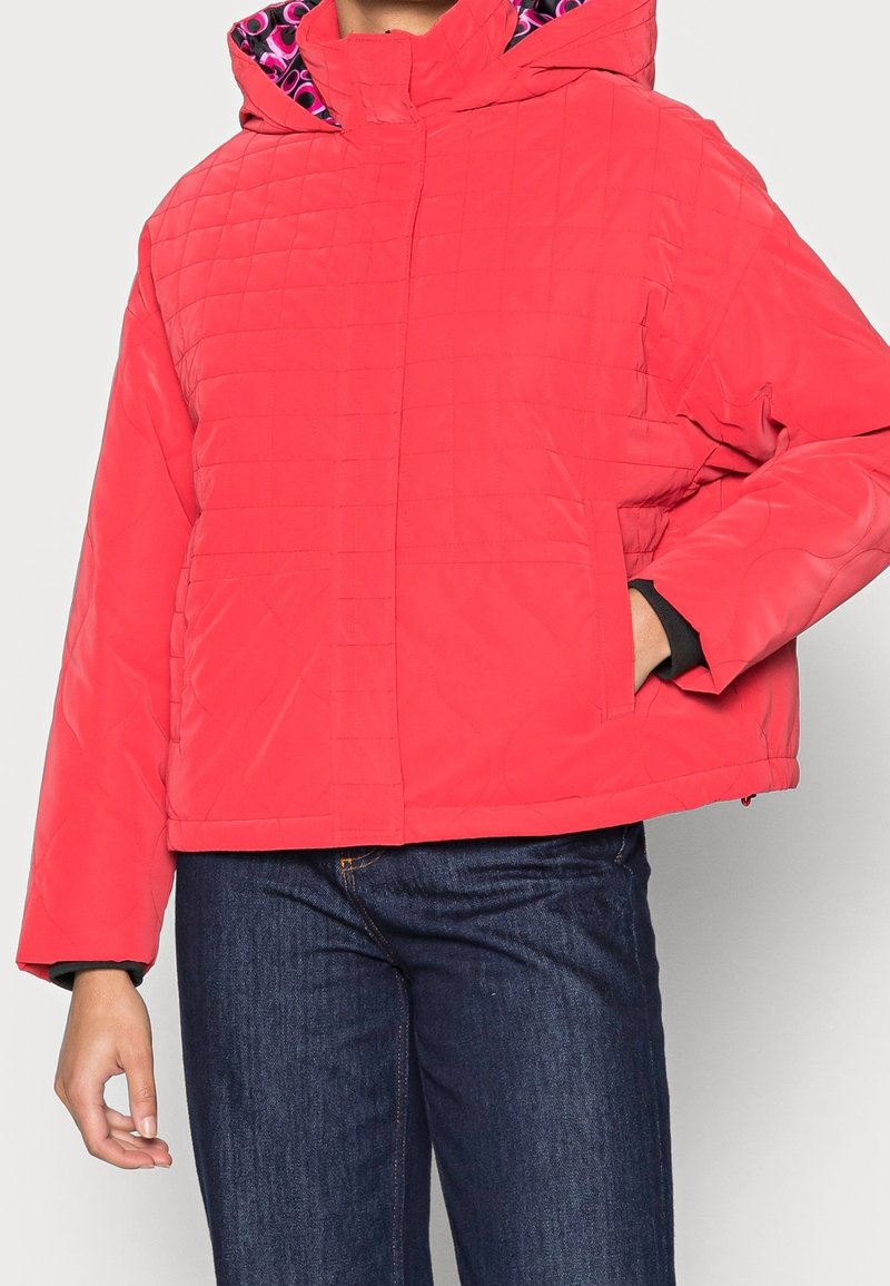 Rød quiltet jakke med foret hætte, der har et pink mønstret indre, lange ærmer og frontlommer, parret med mørkeblå jeans.