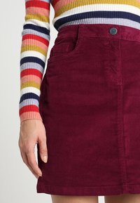 Falda de pana burdeos con un bolsillo delantero, botón visible y remate en el dobladillo. Combinada con una blusa de manga larga a rayas multicolores.