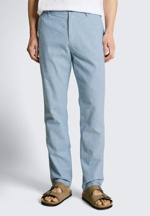 Mężczyzna w jasnoniebieskich spodniach i brązowych sandałach z odkrytymi palcami, stojący na neutralnym tle z rękami swobodnie opuszczonymi wzdłuż ciała.