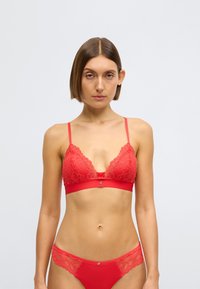 Červená krajková podprsenka s nastavitelnými ramínky, hlubokým výstřihem a jednobarevným červeným páskem. Podrobné krajkové lemování na košíčcích a odpovídající červené kalhotky.