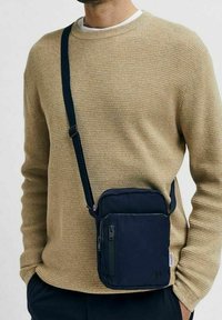 Borsa a tracolla blu navy con tasca anteriore con zip, forma rettangolare, tracolla regolabile e texture morbida, indossata sopra un maglione beige.