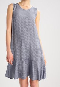 Bezramenné šaty z modré látky se strukturovaným vzorem. Rozšířený střih s volánem na spodním lemu, dosahujícím do poloviny stehna. Hladká textura po celém povrchu.