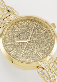Gold-tone watch with a glittery gold dial and crystal hour markers. Features a linked bracelet-style strap adorned with multiple clear stones.