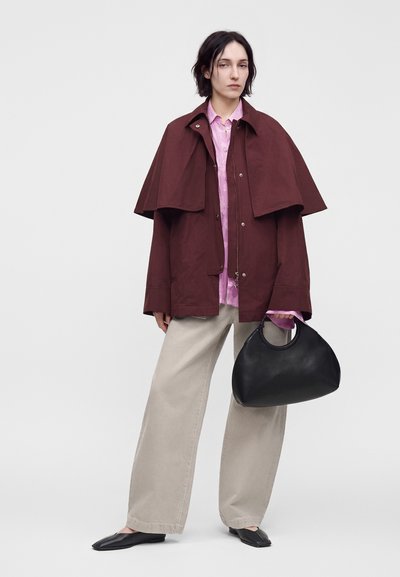 Femme portant une cape bordeaux, une chemise rose, un pantalon beige à jambes larges, des ballerines noires, tenant un grand sac à main noir sur un fond uni.