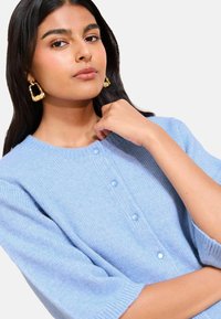 Jasnoniebieski sweter z dzianiny z okrągłym dekoltem, z trzema guzikami i rękawami o długości trzy czwarte. Modelka nosi złote, wyraziste kolczyki.