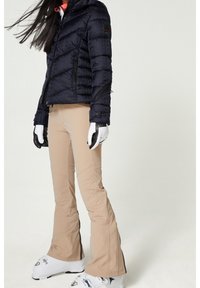 Námořnická prošívaná bunda s vyraženým logem, béžové rozšířené lyžařské kalhoty, bílé lyžařské boty se stříbrnými doplňky a černé rukavice.