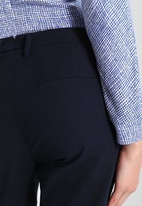 Marinblå byxor med en slät yta och en bakficka. Ovanpå finns en ljusblå mönstrad skjorta med ett rutmönster.