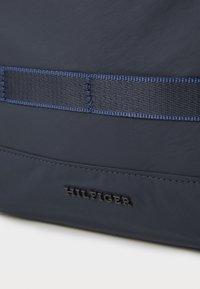 Marineblå stoffveske med glatt tekstur, med en horisontal vevd stropp og preget "HILFIGER"-logo nederst.