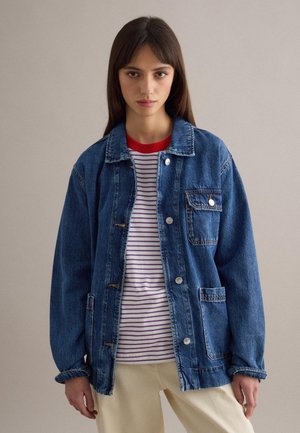 Jeune femme portant une veste en jean bleue par-dessus une chemise rayée blanche et violette à col rouge, debout devant un fond uni.