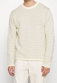 En krämfärgad stickad tröja med ett texturerat mönster av små färgade trianglar i orange och blått. Ribbad halsringning och manschetter. Regular fit.