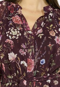 Abito con stampa floreale in un profondo bordeaux, con fiori rosa e bianchi, un'apertura con bottoni e una vita legata per una silhouette definita.
