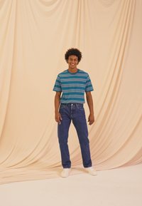 T-shirt a maniche corte a strisce color turchese e blu, jeans blu a vita alta e sneakers bianche su uno sfondo beige chiaro.