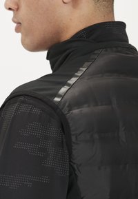 Mustat pehmustetut liivit, joissa on korkea kaulus, teksturoidusta kankaasta ja tyylikkäistä heijastavista raidoista olkapäillä.