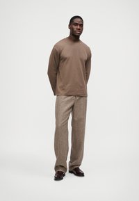 Muškarac stoji u smešnom sme�đem dugim rukavima, teksturiranim bež hlačama i tamnosmeđim mokasinkama pred jednostavnom belom pozadinom.