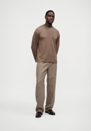 Muž stojaci v hnedej košeli s dlhým rukávom, textúrovaných béžových nohaviciach a tmavohnedých mokasínach na bielom pozadí.