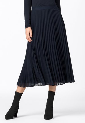 Femme portant une jupe midi plissée bleu marine, un haut noir à manches longues et des bottines à talons noires, debout devant un fond blanc.