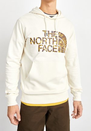 Osoba ubrana w kremową bluzę z kapturem z wzorem logo "The North Face" w kolorze złoto-brązowym, pod nią żółtą koszulkę oraz ciemnobrązowe spodnie.