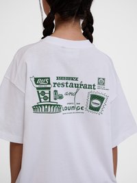 Person med flettehår som har på seg en hvit T-skjorte med grønn grafisk tekst og illustrasjoner som promoterer en stadionrestaurant og lounge.