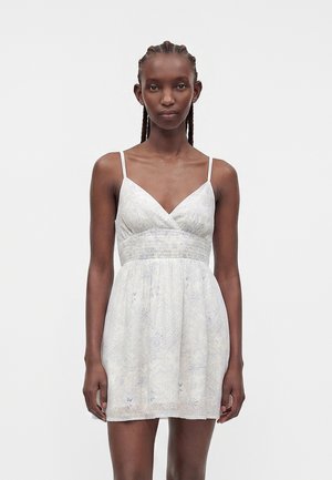 Frau mit geflochtenem Haar, die ein ärmelloses, weiß gemustertes Sommerkleid mit V-Ausschnitt und tailliertem Bund trägt, vor einfarbigem Hintergrund stehend.