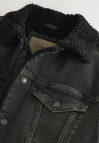 Črna jeans jakna z puhastim črnim ovratnikom, ki ima gumbne zap closures, žep na prsih in teksturirano površino. Oznaka je vidna znotraj ovratnika.