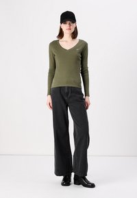 Olivengrønn langermet V-hals topp og svarte vide jeans. Modellen har på seg en svart cap og svarte ankelstøvler, står mot en ensfarget bakgrunn.