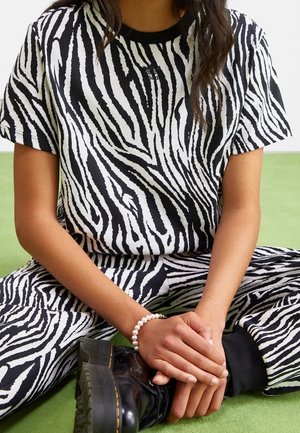 Personne portant une chemise et un pantalon rayés noir et blanc façon zèbre, des bottes noires et un bracelet de perles blanches, assise en tailleur sur un sol vert.