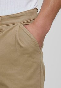Pantaloni din bumbac de culoare bej deschis, cu buzunare laterale, având o textură fină și un detaliu subtil de cusătură pe margini.