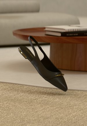 Chaussure plate slingback en cuir noir à bout pointu avec un accent doré sur le bout et le talon, placée près d'une table basse ronde en bois et d'un tapis beige.