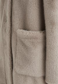 Helles beiges Kunstpelzkleidungsstück mit einer weichen, flauschigen Textur und einer sichtbaren Tasche auf der Vorderseite. Der Stoff hat ein glattes, plüschiges Aussehen.