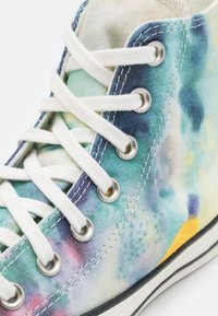 Close-up af højtoppe sneakers med flerfarvet tie-dye stof, hvide snørebånd trådet gennem metaløjer, og hvid gummisål.