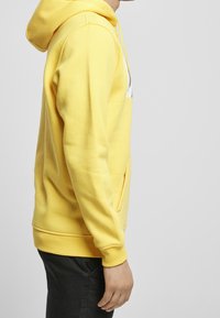 Žlutá mikina s kapucí, kangaroo kapsou, žebrovanými manžetami a lemem. Obsahuje bílé logo na hrudi a hladký povrch.