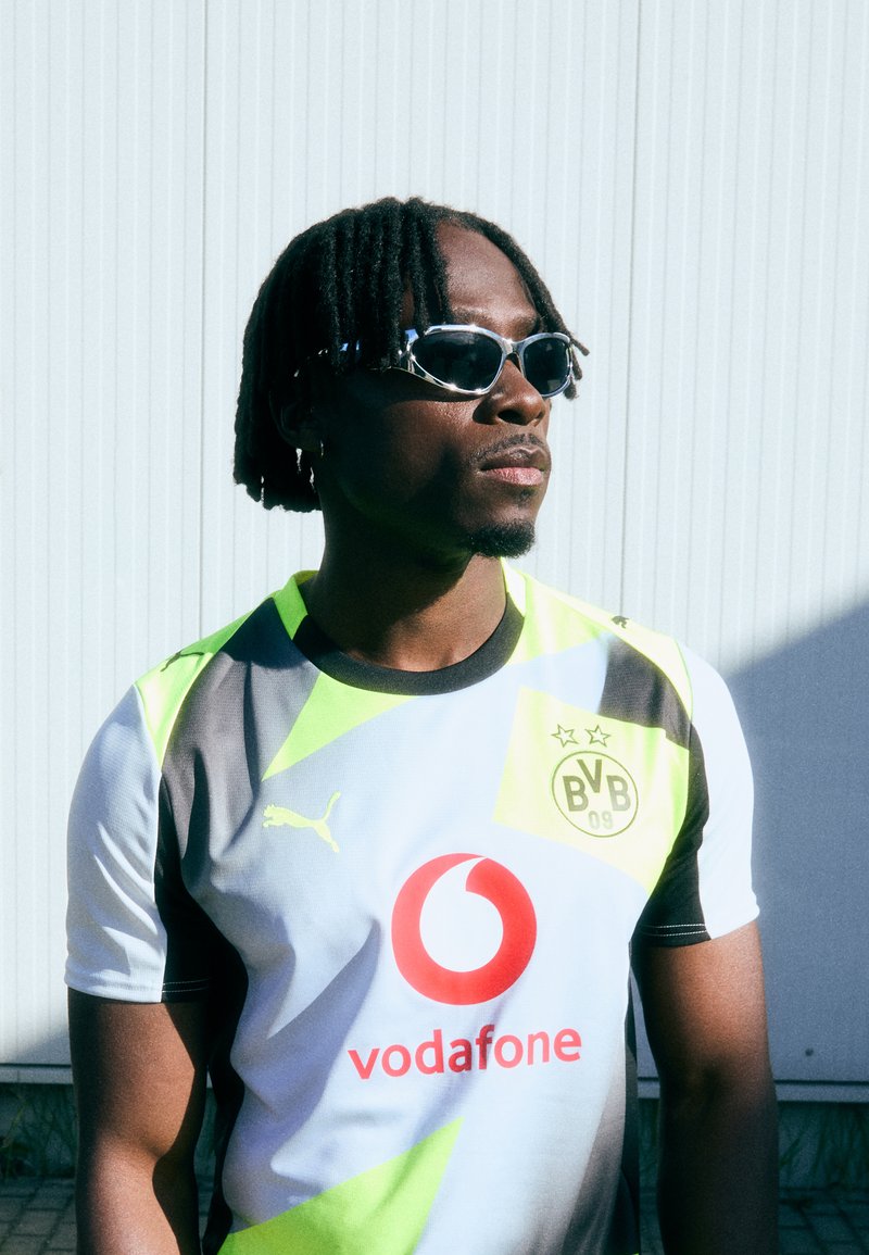 Maglia caratterizzata da colori nero, bianco e verde neon con motivi geometrici. Include un logo Vodafone e un marchio Puma. Accessori: occhiali da sole.